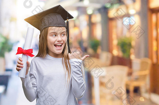 年轻漂亮的女孩戴着毕业帽，在孤立的背景上拿着学位，用拇指指向一边，脸上带着开心的微笑