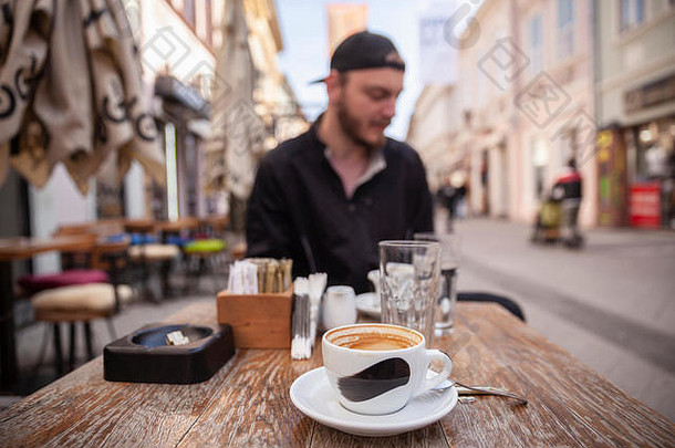 咖啡杯特写，用刷子在杯子上印上<strong>粗线条</strong>。坐在桌子上，在街上的户外咖啡馆里。坐在桌子对面的那个人。行人