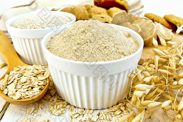 面粉和麸皮燕麦放在白碗里，燕麦片放在汤匙里，燕麦茎，面包和饼干，餐巾放在木制的背景上