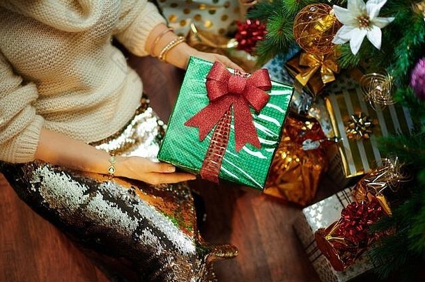 特写镜头：在装饰过的圣诞树下，一位身穿金色亮片裙和白色毛衣的年轻女子在礼物盒旁展示圣诞礼物盒。