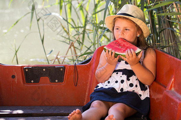 一个穿着裙子戴着帽子的可爱小女孩坐在湖边的船上吃着多汁的西瓜。她喜欢在自然中度过时光
