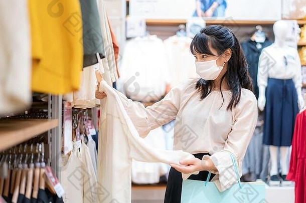 一名亚洲妇女戴着口罩在脸上，在购物中心挑选带有购物袋的衬衫，用于保健和预防冠状病毒（Covid19）