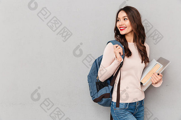 一个聪明的女人背着书包在大学里学习，拿着书和笔记本电脑在灰色的<strong>墙</strong>上