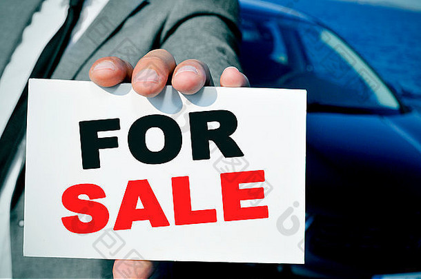 一名西装革履的男子手持一块写有待售字样的招牌，背景是一辆黑色汽车