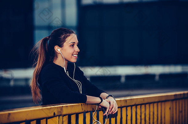 穿着黑色运动服的年轻女子，晚上在城里的桥上跑步后，一边听音乐一边休息。