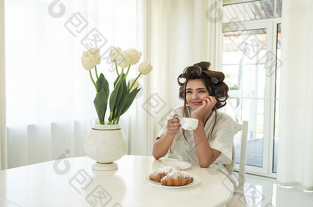 早上在明亮的厨房里，戴着耳机，戴着卷发器，端着一杯咖啡，吃着牛角面包的年轻漂亮黑发女郎