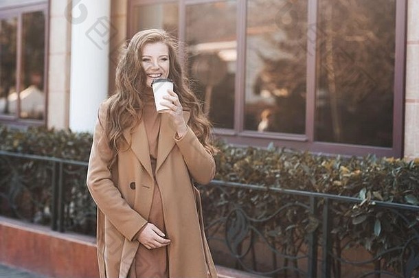 在阳光下的特写镜头中，一位快乐的孕妇穿着时尚的米色外套和连衣裙，从外卖杯里喝咖啡，漫步在城市街道上。春夏