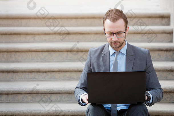 年轻的专业人士坐在公园的台阶上，双手放在笔记本电脑上。这位商人看着笔记本电脑屏幕，非常满意