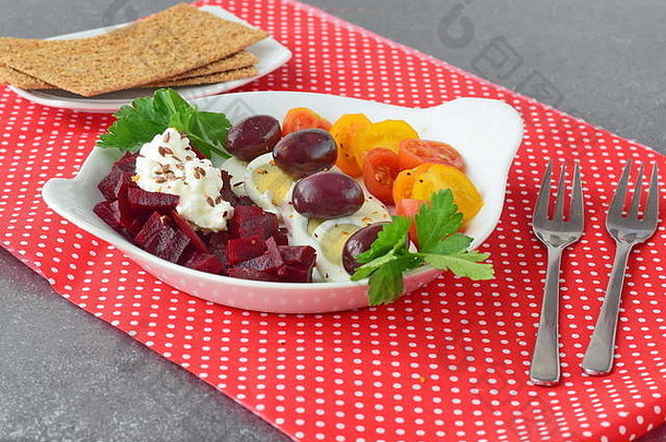 简单早餐：甜菜根、樱桃番茄、鸡蛋、白干酪和薯片放在一个白色盘子里，背景是红色的纺织品。健康食品概念。