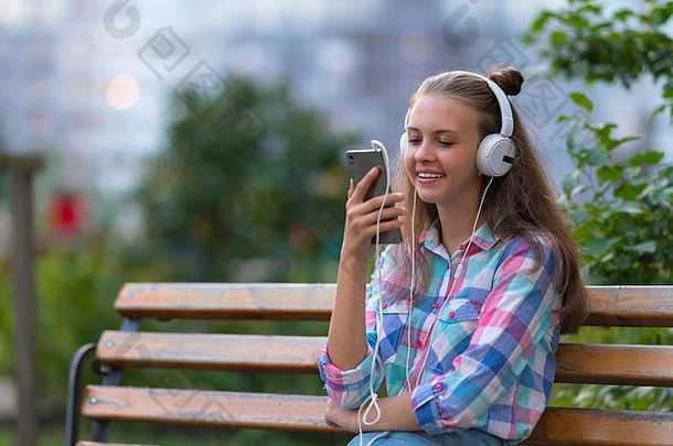 快乐的年轻女子在花园或公园的长椅上放松地听着音乐，带着愉快的微笑和复印空间