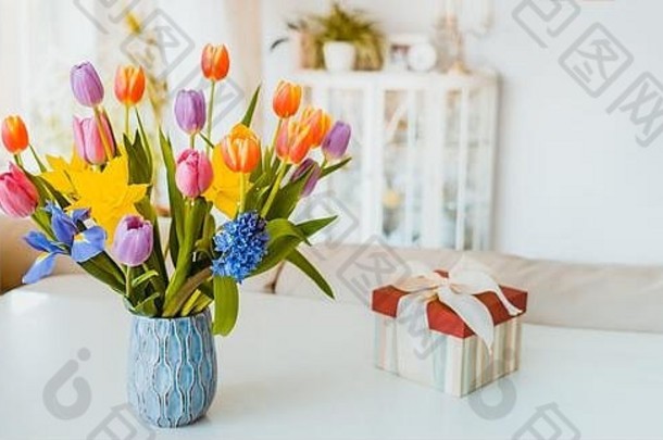 清新的春天，在白色的桌子上，有郁金香、水仙花、鸢尾花和礼盒，背景为浅色经典设计的房间。节日人流