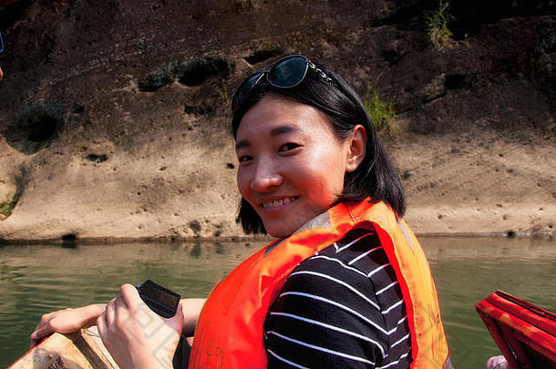 中国福建省武夷山九曲河畔，一名身穿橙色救生衣、面带微笑的中国女子坐在木筏上。