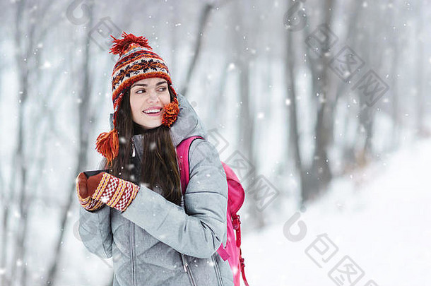 穿着灰色夹克、帽子和连指手套的旅游黑发女孩手持<strong>保暖</strong>杯站着，在寒冷的<strong>冬季</strong>森林里思考<strong>冬季</strong>风景
