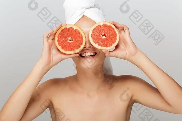 没有化妆的微笑女孩的肖像，头部用白毛巾遮住眼睛，灰色背景上有一半葡萄柚