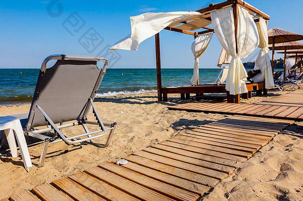 木制小径穿过沙滩，通向舒适的木制凉亭，凉亭上有白色窗帘，阳光明媚的海滩。