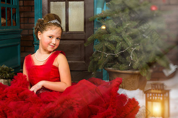 身着红色礼服、戴着珍贵皇冠的冬季小公主在迷人的假日室内迎接新年和圣诞节