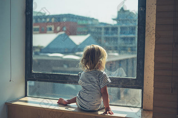 一个蹒跚学步的小孩正坐在一座城市公寓的窗边向外看