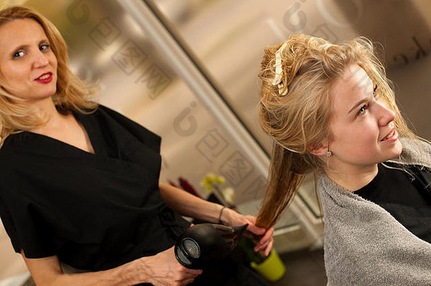 工作中的专业发型师-在专业工作室为年轻漂亮的金发顾客做发型的理发师