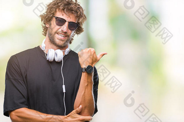 英俊的西班牙裔男子戴着耳机在孤立的背景下听音乐，面带笑容，面带笑容，并用手指着thu的一边