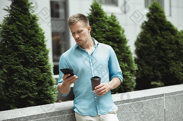 这位年轻英俊的男子站在办公商务中心附近，边走边喝咖啡，一边检查他的智能手机