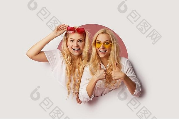 两个戴着心形太阳镜的年轻快乐、震惊的女人的肖像，透过墙上的白色孔洞望去。大减价。滑稽的面孔。空位