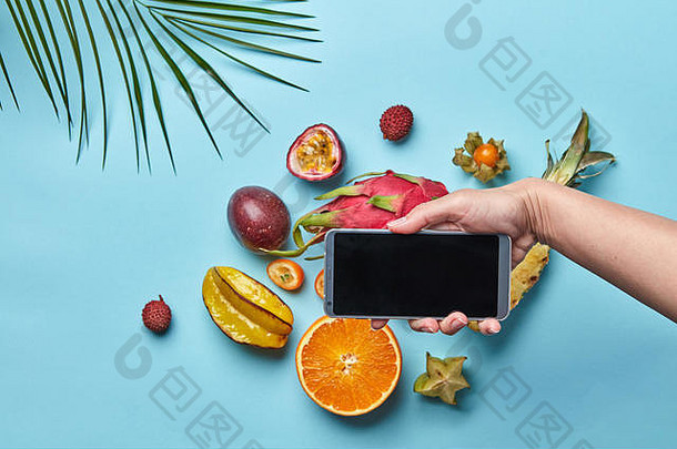 一组不同的热带水果和棕榈叶放在一个蓝色音标上，并留有文本空间。女人的手拿着手机。在线购买产品的概念。平铺