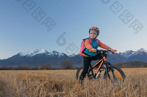 一个白人孩子在麦田里骑自行车。小女孩在美丽的雪山背景下走着<strong>黑橙</strong>色的自行车。自行车看台