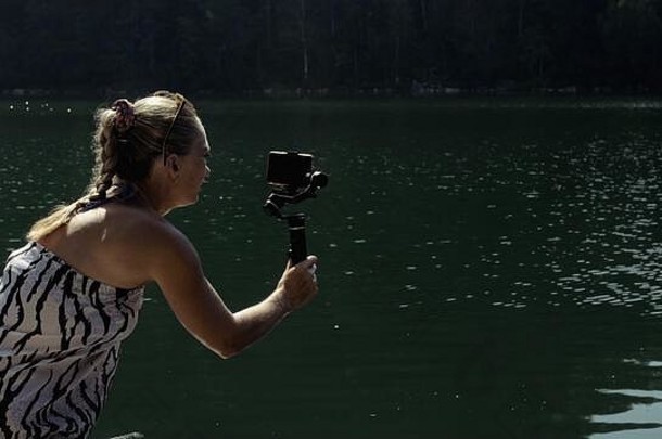 女子在手持胶片上拍摄智能手机万向节稳定装置。女孩站在码头上自拍。博主播放视频博客。