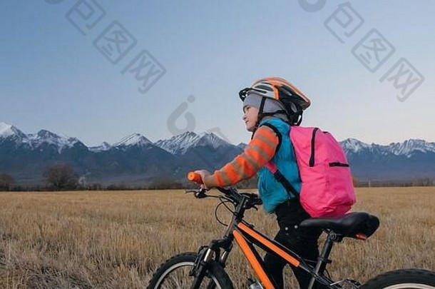 一个白人孩子在麦田里骑自行车。小女孩在美丽的雪山背景下走着黑橙色的自行车。自行车看台