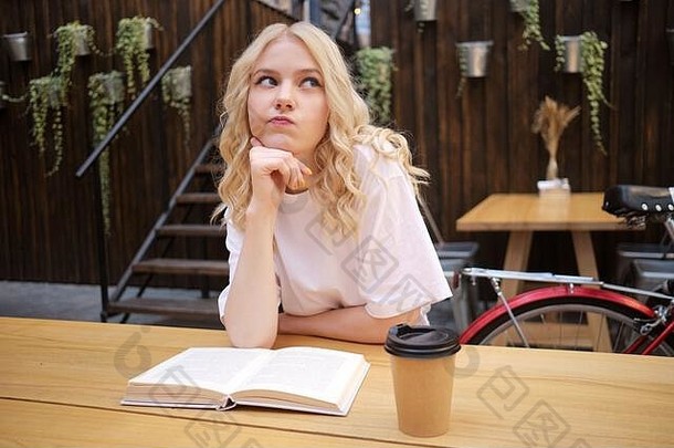 在城市咖啡馆的院子里，一个迷人的金发女孩带着书和咖啡若有所思地看着别处