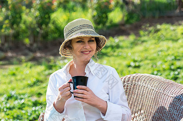 戴帽子的女人坐在花园里，手里拿着<strong>一杯咖啡</strong>