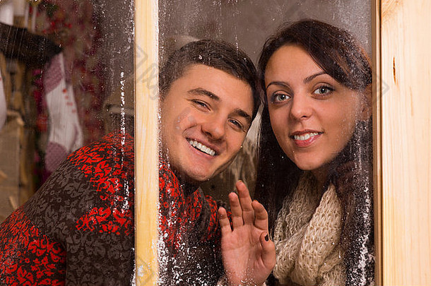 特写镜头：穿着冬季长袖服装的年轻情侣们微笑着站在玻璃窗后面，看着相机。