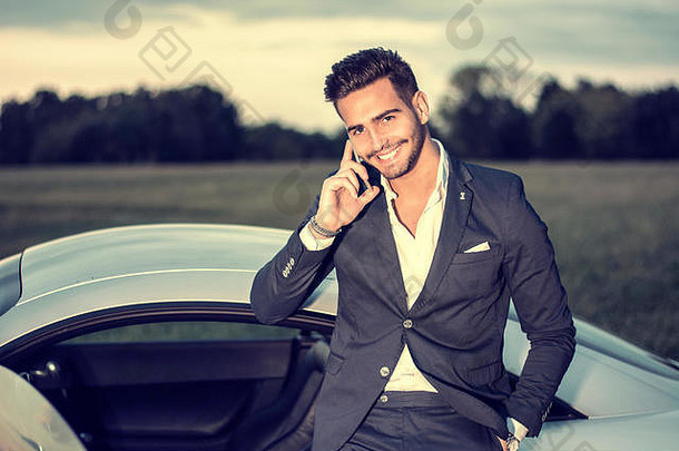 穿着西装坐在他新的时尚汽车里的年轻魅力男子的肖像，在乡村户外