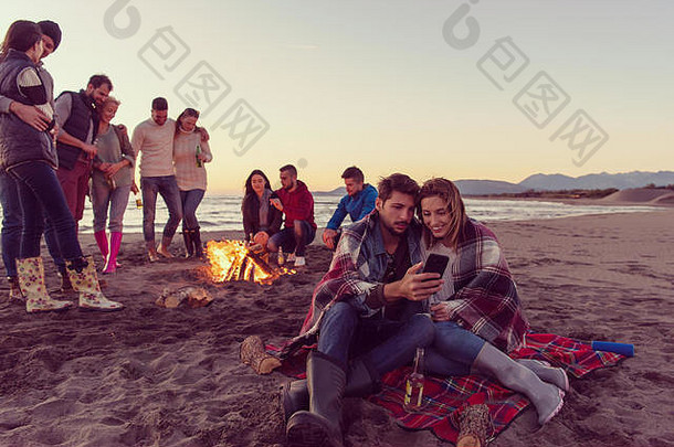 这对夫妇在秋季海滩派对上使用手机，和朋友一起喝啤酒，玩得很开心