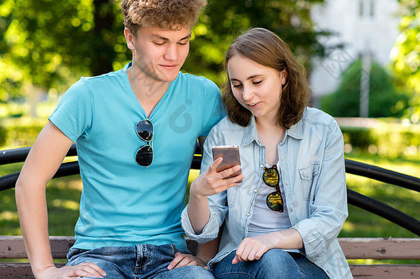 一男一女坐在长凳上。<strong>大</strong>自然中的夏天。手里拿着智能手机的女孩。情绪化的微笑可以<strong>解读</strong>社交网络。