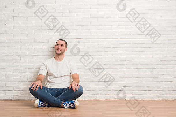 年轻的白种<strong>人</strong>坐在白砖墙上方的地板上，微笑着向一边看，凝视着别处思考。