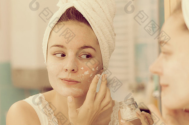 女人在脸上涂面膜保湿护肤霜，对着浴室镜子看。呵护肤色的女孩分层膏