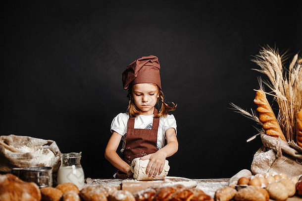穿着围裙，戴着帽子的迷人小女孩站在桌旁揉面包面团，享受烹饪的乐趣