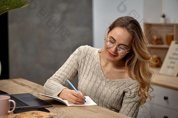 金发女郎女孩博主眼镜米色毛衣微笑坐着厨房木表格写笔记本关闭