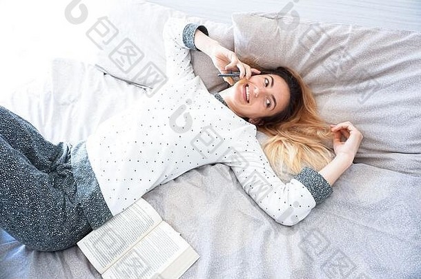 一个女孩穿着睡衣呆在卧室里，一边用智能手机打电话，一边在床上<strong>翻开书本</strong>——在家里放松地呆在家里