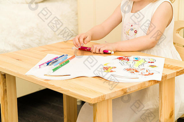 活泼的学龄前儿童，可爱的蹒跚学步的金发女孩，用彩色铅笔和毛笔在纸上画画，坐在家里或幼儿园的木桌旁