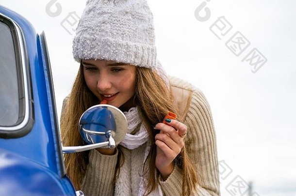 美丽的年轻女孩看着汽车后视镜，撅起嘴唇。<strong>秋冬</strong>时<strong>节</strong>，在蓝色复古汽车旁戴着帽子和围巾、留着长发的女人，模仿