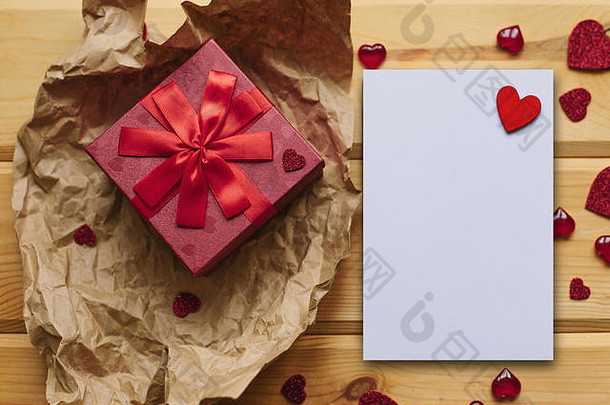红色的盒子里有一件礼物，旁边是一张白色的空白纸，上面有一个心形的木纹。情人节、妇女节或母亲节的节日概念。