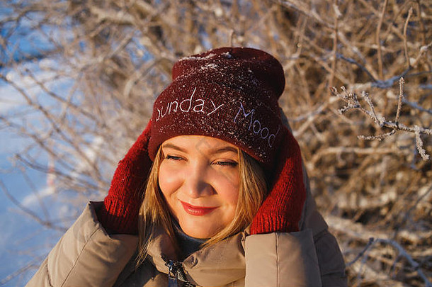 美丽的年轻白人妇女，戴着红色手套和红色冬帽，在雪地里享受着阳光明媚的冬日。她拿着帽子望着太阳。好心情、好天气、好冬天的爱情观念。