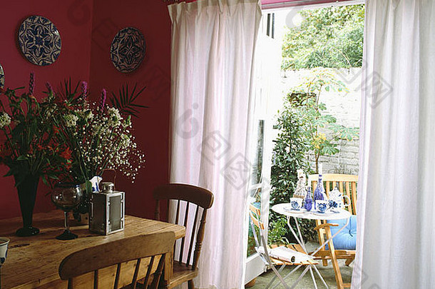 粉色餐厅中的松木桌椅，法式门上有白色窗帘，可以看到天井花园