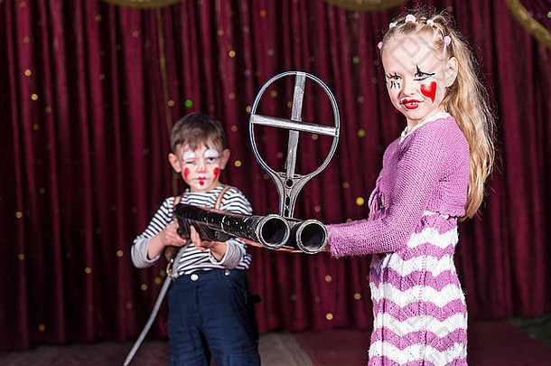 男孩和女孩打扮成小丑站在舞台上，红色的窗帘上拿着一把双管大炮，铁制瞄准镜