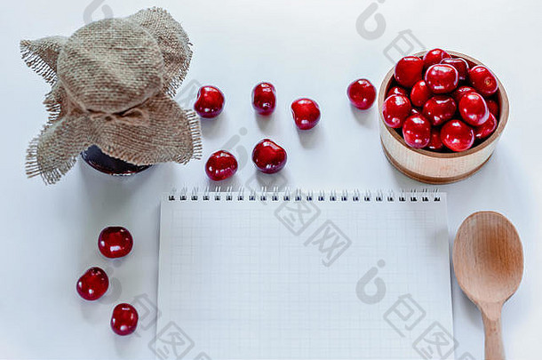 红色成熟樱桃配果酱罐、木勺和白色背景的笔记本。平铺。食物概念。
