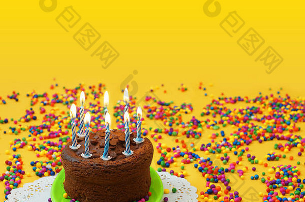 巧克力生日蛋糕，蓝色和白色蜡烛点燃，放在一个绿色小盘上，周围是散布在黄色背景上的糖果球