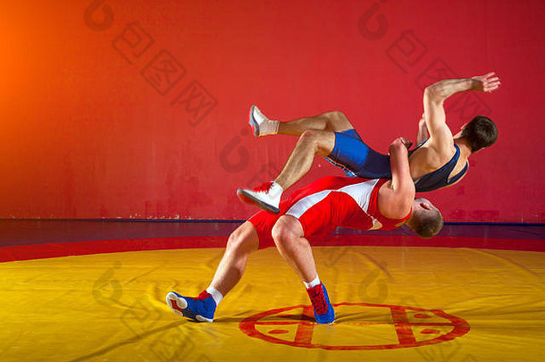 两名身穿红蓝制服的希腊罗马摔跤手在体育馆的黄色摔跤地毯上进行背景摔跤