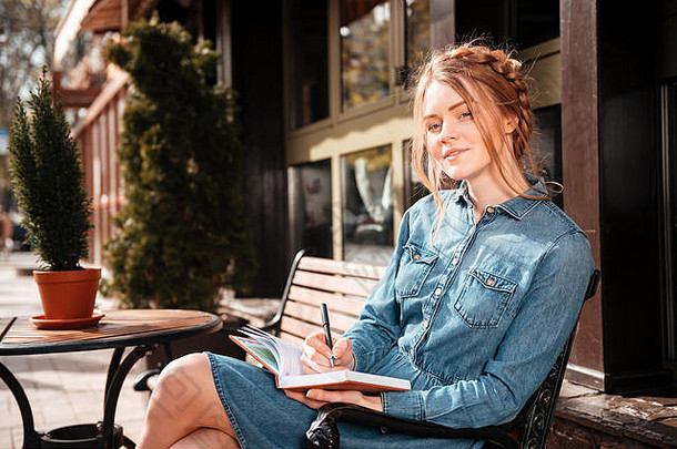 坐在露天咖啡馆里，一位沉思的可爱年轻女子在笔记本上休息和写作